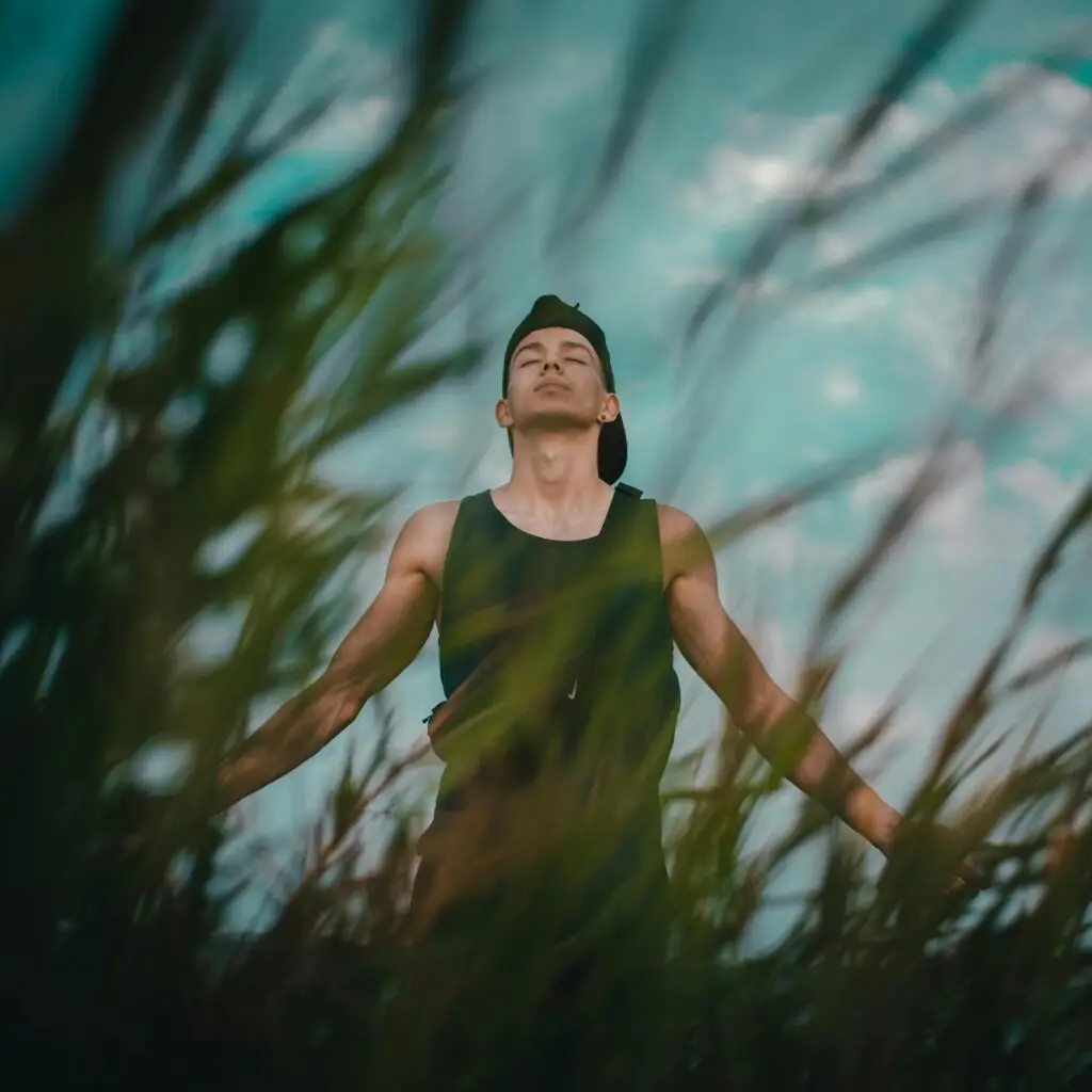A young man relaxes and meditates among tall grass outdoors, eyes closed, embracing nature.